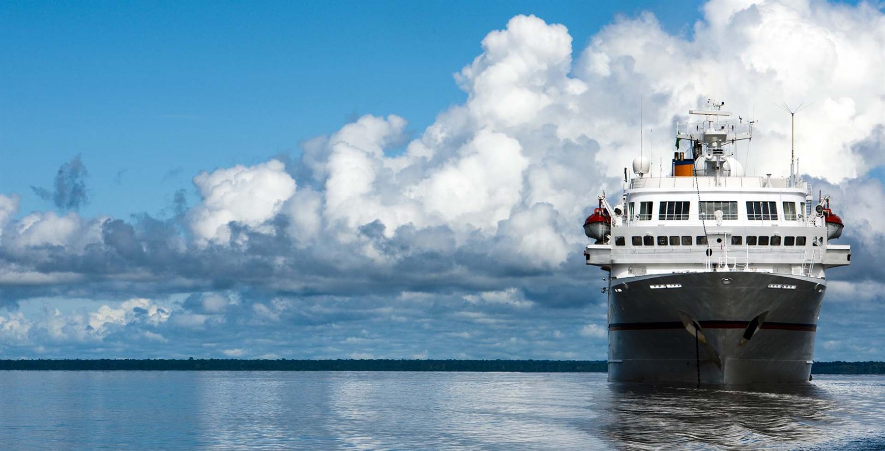 Small expedition cruise ship on a wide river with a line of rainforest on the horizon
